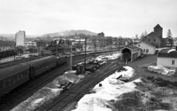 Asker stasjon fotografert omkring 1970. Til høyre ruver transformatorbygningen fra 1922. På den andre siden av sporområdet skimtes den nye stasjonsbygningen fra slutten av 1950-årene og man ser Fusdalgården. I bakgrunnen ses Rådhuset og i det fjerne skimtes Asker kirke. Foto: Ukjent / Budstikka