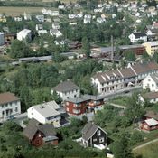 Bortre husrekke langs Strømsveien fra v.: Apotekgården, Farupgården, Wiborg & Gulbrandsen, Farvehandler Sørensen og Strømmen Melkeforsyning. Hitterste rekke Kjustadgården med postkontoret fra 1948, rødt hus dr. Frey. Strømmen Trevarefabrikk og Høvleri i bakgrunnen. 1960.