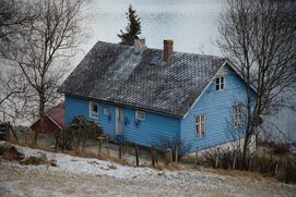 Eit blått hus ved fjorden. På taket er det to ulike pipar. Langs den eine veggen står ein liten vedstabel. Eit lavt gjerde står mellom kameraet og huset.