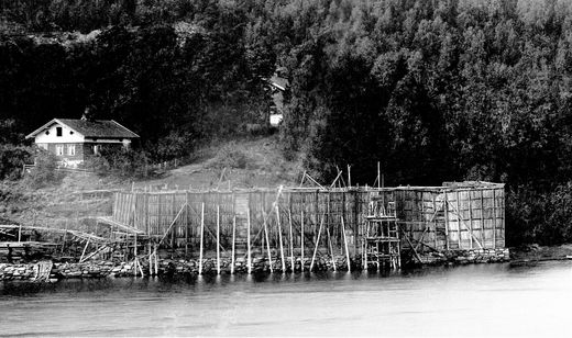ISSTABEL VED BRYGGEBAKKEN 1890-TALLET. Lastebåtene/seilskutene la seg inntil isstablen og lastet isen rett om bord. Midt på stabelen er det tilrettelagt for å åpne den og ta ut isen i flere plan. Eiendommen bak stabelen er Bryggebakken. Delvis skjult av trærne ligger Syverstadgården. Denne isstabelen var ikke lenger i bruk etter 1. verdenskrig. Utsnitt av større fotografi. (Foto fra AS Søndre Nærsnes)