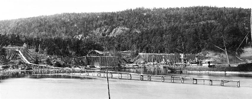 ISSTABLER OG ISHUS PÅ 1890-TALLET. Dette er fra den tiden da det ble produsert mest is på Nærsnes. Helt til venstre ser vi isstabelen på Mellomstranda, der Nærsnes Kystlag nå har en liten replika av en isstabel. På bildet ser vi at den permanente lange isbrygga (isrenna) har blitt anlagt. På Flatstranda er det nå kun ett ishus igjen. 3 store isstabler har estattet de andre ishusene og økt den samlede lagringskapasitet. Lastebåten ligger ved det som senere ble kalt "Den store isbrygga". Detaljer i bildet viser isblokker på vei ut til skipet. Brygga ble revet på 1970-tallet. Restene av grunnmuren av isstabelen foran ishuset ligger fremdeles i strandkanten, der kyststien passerer over. Utsnitt av et større fotografi (Foto fra AS Søndre Nærsnes, fotograf ukjent)
