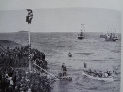 De kongelige kom til Harstad onsdag 31. juli 1907. Byen hadde kai som kunne ta imot D/S «Olav Kyrre», som for anledningen var kongeskip, men en pågående konflikt med kaieier Anna Elisabeth Kaarbø gjorde at kommunen valgte en provisorisk løsning på Hamnneset. Det var ordfører Ivar Lund og magistrat Carl Dahl som tok imot kongefølget ved Tjuvholmen. De var de første som ble rodd i land og det er kanskje de to vi ser på den provisoriske brygga for å ta imot kongeparet. Avisa Haalogaland fortalte at det blåste nordøstlig kuling. Også Fridtjof Nansen var med på kongeferden, men avisen nevnte ikke hans navn. Fra Simensen, Sigurd: Harstad gjennem 50 år, Harstad 1953.