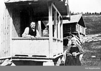 Bilete 3: Tolleiv og Mari, med strikketøyet, utanfor Søre Juvsjordet, dit dei flutte i 1909, etter at sonen Elling overtok Tufto. Mari i stakk med ruta skjorte og plagg på hovudet. Ukjent fotograf, Hol bygdearkiv, 1910.