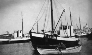 M/S «Vidar» H-37-AM, byggeår 1915, 37 fot lang, motor 20 HK Søderblom. Eigar Jakob Wikene. Han kosta den første motorbåten på Fedje, truleg i 1903. Det var ei lita snekke med 2 HK oppfyringsmotor. Dette var innleiinga til ei gradvis utskifting av dei tradisjonelle ro/seglbåtane med båtar med motor som drivkraft. Denne utviklinga skaut verkeleg fart under den første verdskrigen. Ifølgje merkeregisteret i 1922 hadde omtrent alle fiskebåtane motor. Dette gjorde sjølvsagt fisket mindre slitsamt. – På bildet sit Asbjørn Mongstad i robåten, medan Ingvar Berg til venstre står og malar.