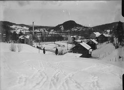 Hamang-området sett fra fra Bjørnegård-området, med Fabriken Tomten fra 1912 til venstre for mennene i bildet, og med Hamang Papirfabrikk fra 1907 bak denne. Sandvikselva bak trærne som går gjennom bildet. Bebyggelsen på denne siden av elva ligger til veien som i 1955 fikk navnet Industriveien. Foto: Narve Skarpmoen/Nasjonalbiblioteket (1912–1930).