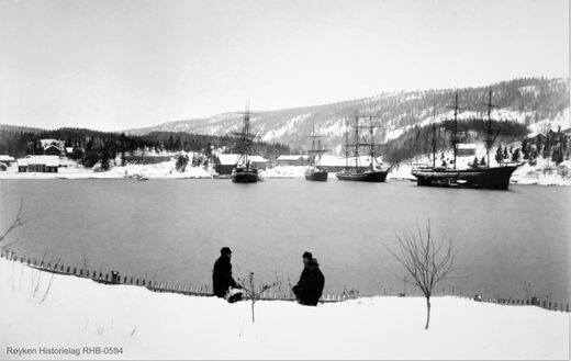 SEILSKUTER I NÆRSNESBUKTA 1880-TALLET. Bildet tatt fra Saltbutangen/Steinborg. Husene til eiendommen Nærsnesstranden til venstre. Mellom husene og skutene er Isstablen på Mellomstranda. Brygger for lasting av is er ennå ikke lagt ut. Bak skutene sees flere permanente ishus med saltak. Disse ble senere revet og erstattet av åpne isstabler. En av skutene ligger høyt i vannet. Den har losset ballasstein, men ennå ikke fått ombord is.(Foto fra Røyken Historielag, fotograf ukjent)
